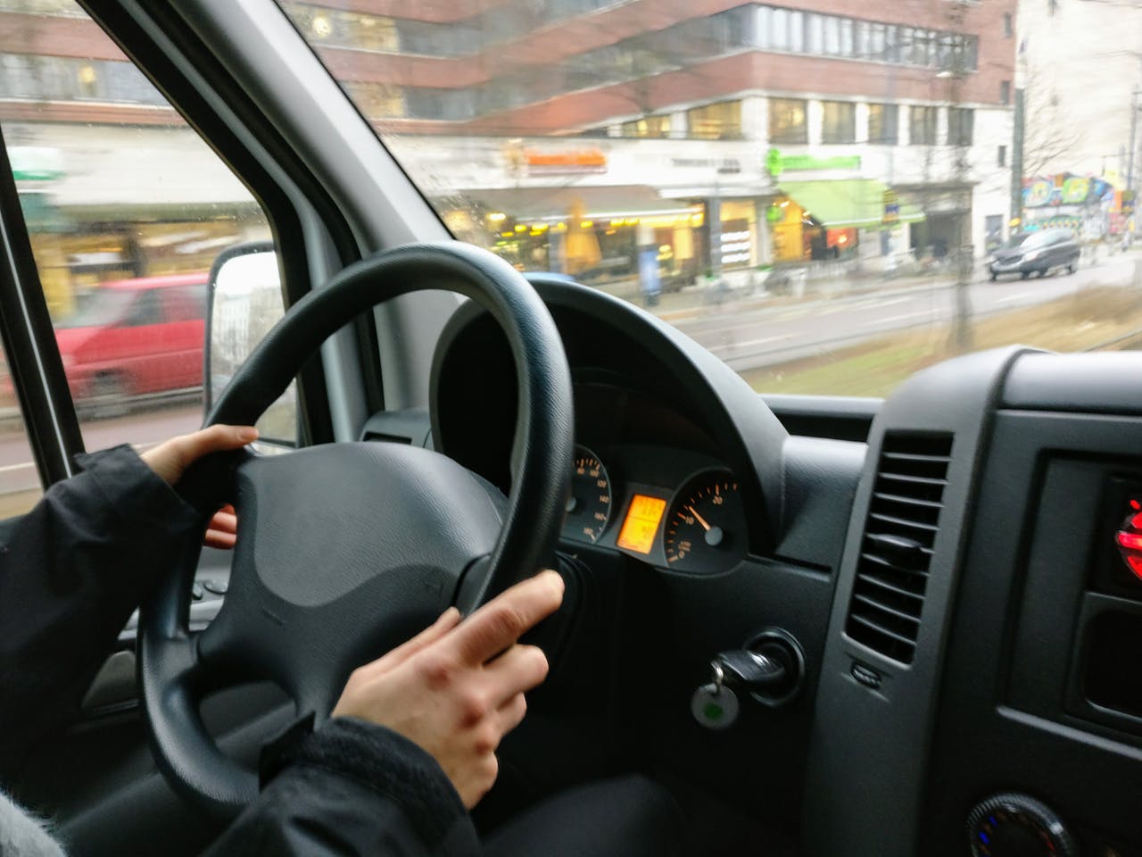 about-us-01 Hands on a steering wheel driving a van through an urban area with shops visible outside.